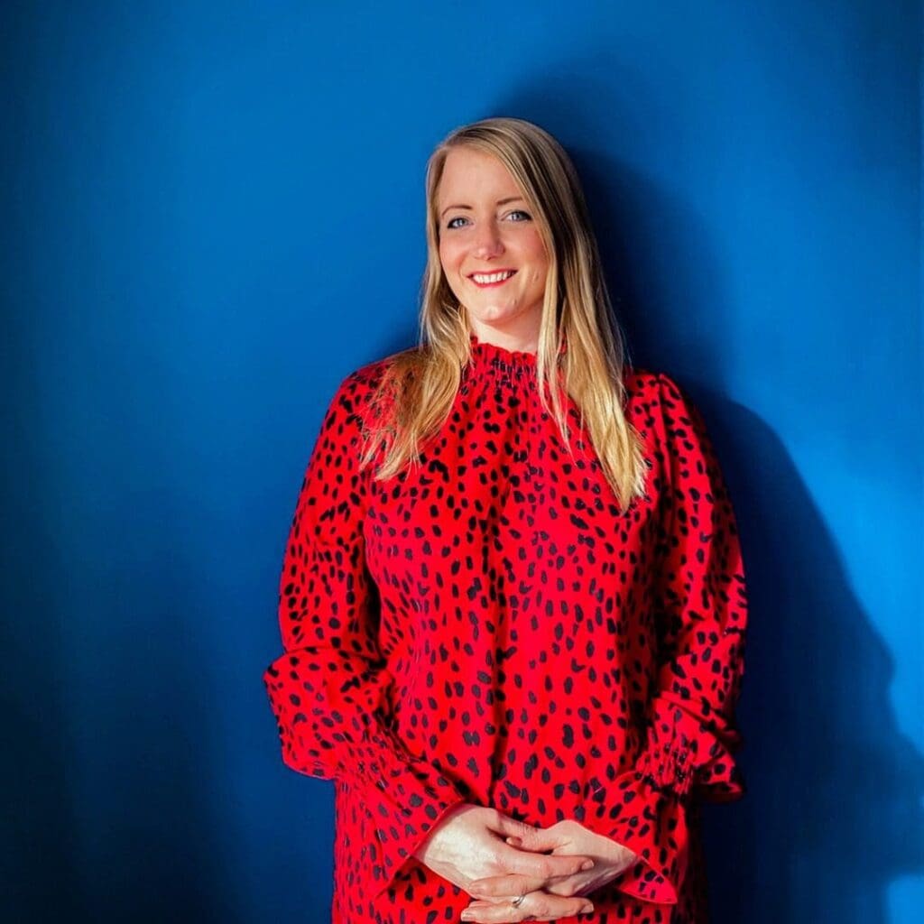 Blue background. White woman in 40s ssmiling long blond hair red long sleev dress black spots hand together below tummy