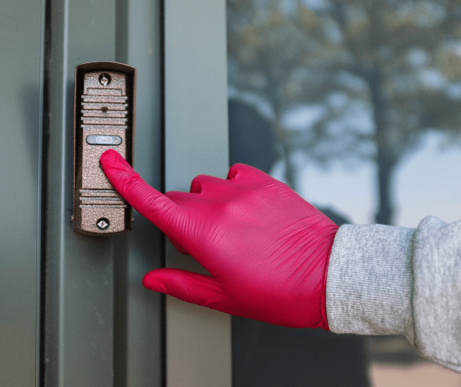 Blurred treest in background, hand in latex pink glove and grey cuff ringing door bell/intercom on vertical slatted green wood.
