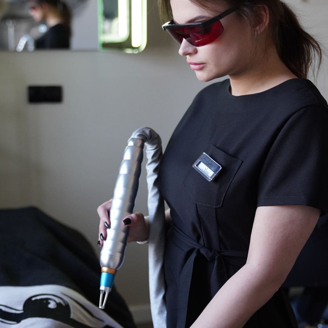 Woman in blue medical top short sleeves and dark glasses. Brown hair tied back. White woman. Holding long tube with laser removal at and looking at tattoo on a back of person lying down