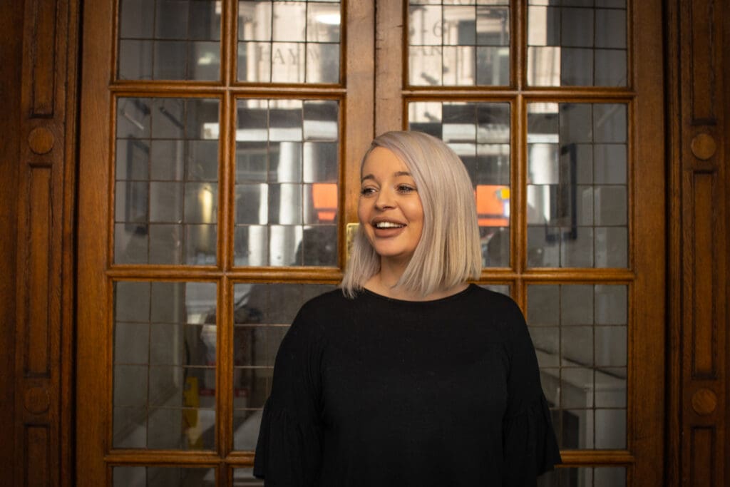 Wona standing in front of glass doors with wooden frames. She is looking to the left hand side smiling/laughing shoulder length straight blond hair black jumper