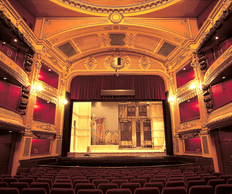 long shot of a stage back of empty red seats balcony either side dark red curtains open on stage with shelves standing to the right