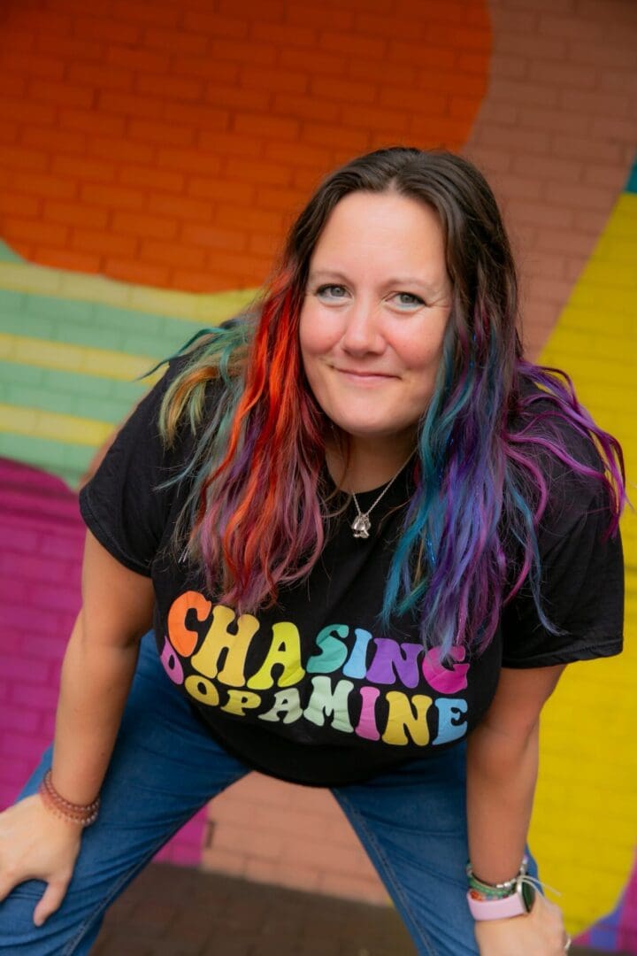 Woman in early 40s long rainbow hair smiling at camera black t shirt with "Chasing Dopamine" in capital letters rainbow colours