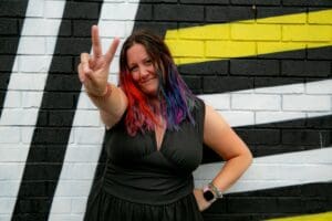 White woman in early 40s in front of brick wall painted white with black horizontal stripes with yellow inbetween top two black lines to the left two black vertical diagonal stripes, Long straight rainbow hair hand on hip other arm out stretched making victory sign smiling in black sleeveless top