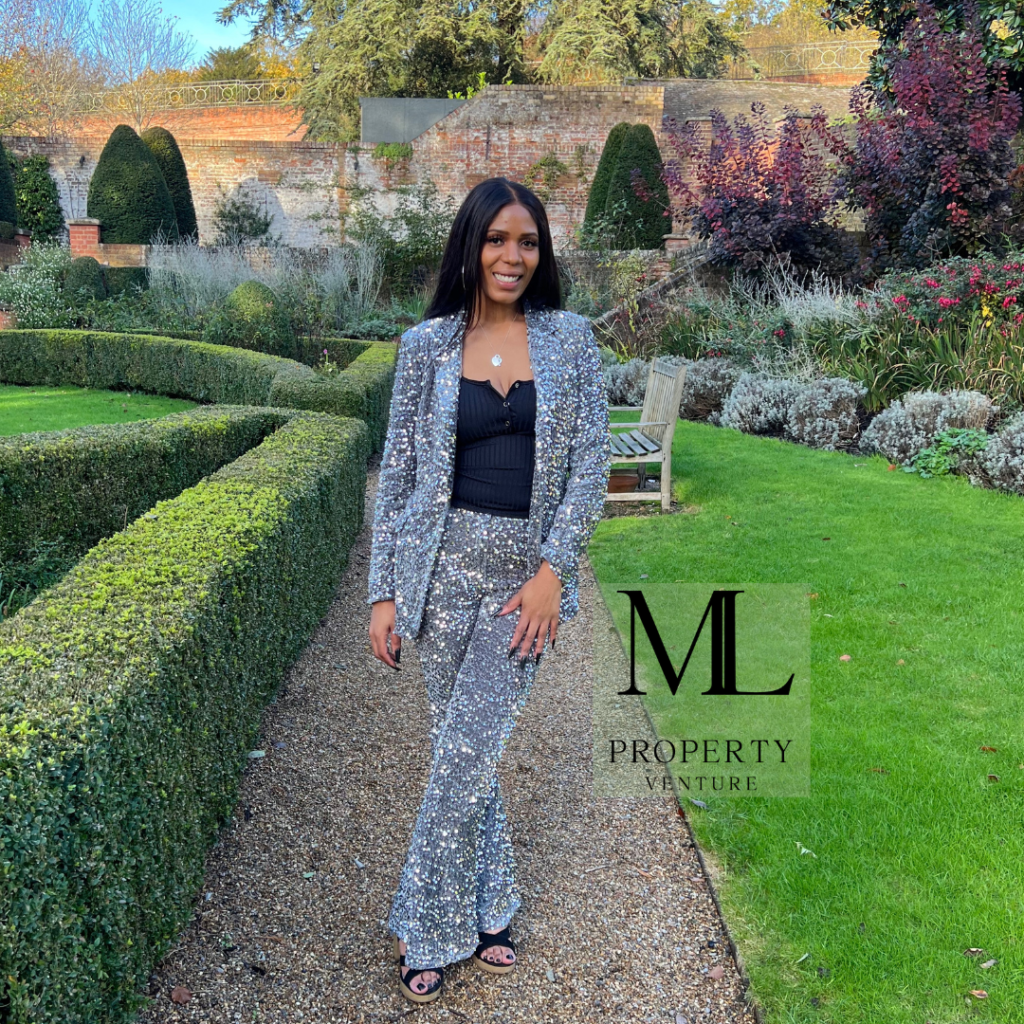 Black woman in black and white trouser matching jacket black top. Left leg crossed over right. Smiling in courtyard green grass box hedge to left standing on path. Flower beds behind. Logo to the right
