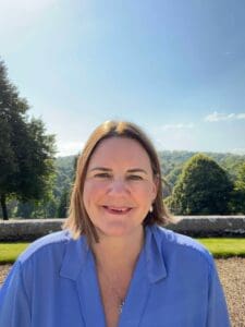 Head and shoulders shot white woman in 40s smiling short brown hair in bob light blue shirt outside in front of green trees and lawn