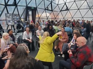 Lots of people sitting down in windowed room, back of person with brihjt yellow jacket arms outstretched