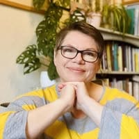 White woman with very short brown straight hair. Glasses, smiling elbows on table hands yellow and grey striped jumper sitting in front of book case with books on it