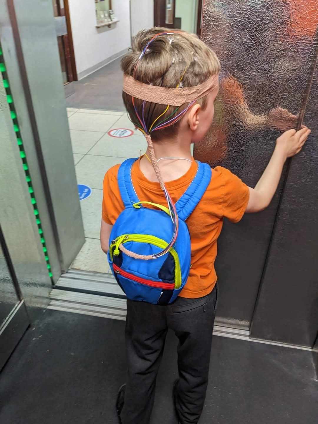 4 year old boy with an EEG with wires stuck to his head and going into a blue backpack facing lift door wearing orange t shirt
