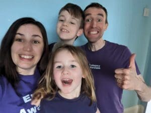 White family all smiling at camera. woman in early 40s at left young girl front middle, behind her young bot man in early 40s to the right all wearing purple t shorts