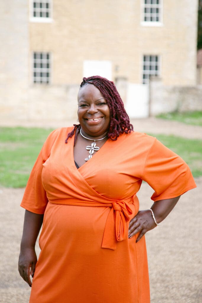 black woman smiling orange dress hand on hip short hair in front of house with grass in front