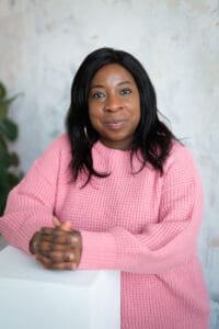 Black woman in late 30s straight shoulder length black hair. Pick loose jumper, hand together on table in front. Smiling head and shoulders