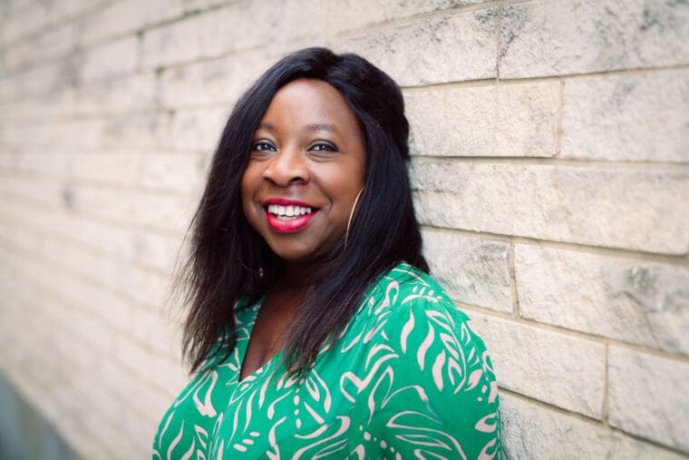 Black woman in late 30s straight shoulder length hair standing in front of wall smiling at camera head and shoulders green dress with white flowers