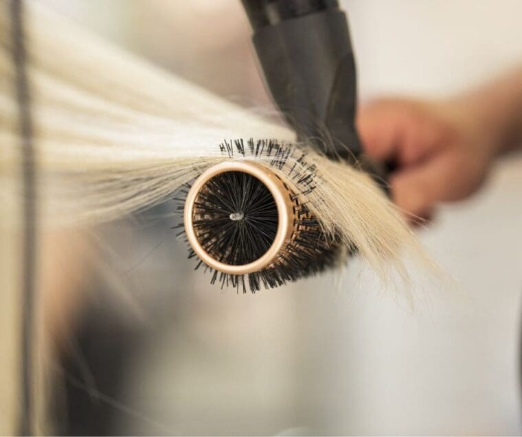 straight blond hair pulled out over round bristled brush white hand holding the brush and end of hairdryer touching the hair on the roller