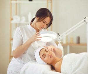 white woman lying on bed towel round head and body see head and shoulders down to middle white woman standing over with facial stand over other woman's face. She's in white uniform