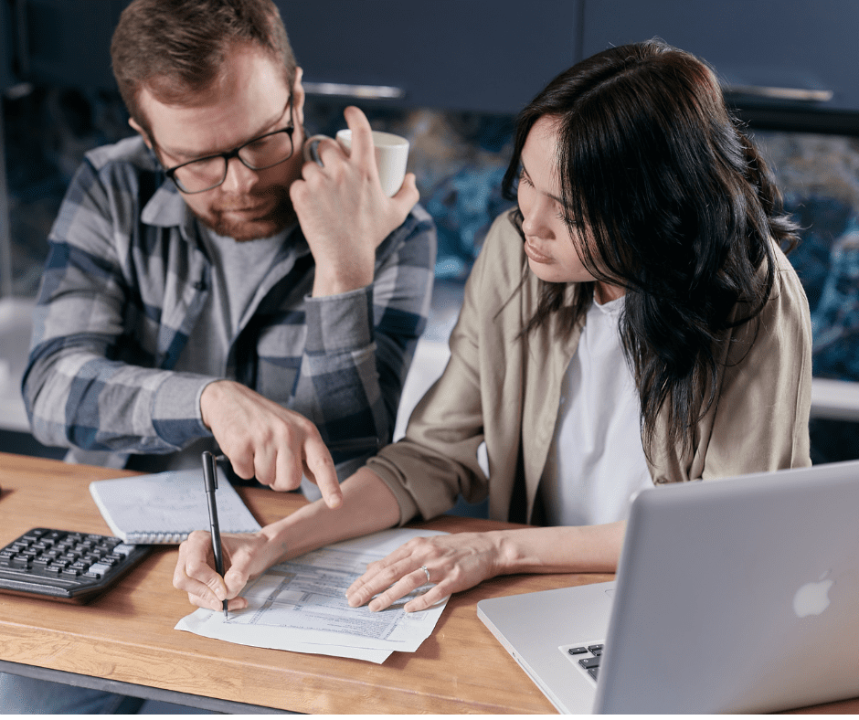 2 people sitting at table with lap top looking at bills. White man in late 20s on left one hand up one hand on the bill pointing, Asian woman in late 20s both hands on the bill pen in in one hand. Lap top open on table to right. Man in blue large checked shirt woman in white top