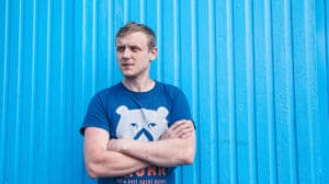 White man in 30s light blue corragated background arms folded blue t shirt white bear on front looking to the left