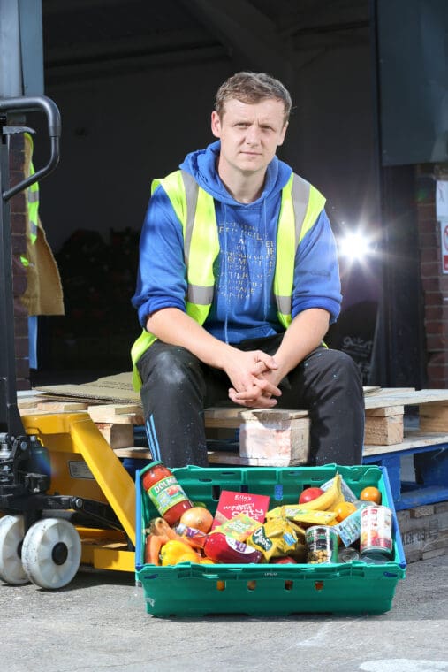 White man in 30s sitting down black background looking at camera. Blue shirt rolled up sleeves florescent sleeveless tello, black jeans hands together in front, supermarket crate of food in front