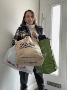 Woman in late 30s standing by front door with large carrier bags front one brown paper bag with Boots on it