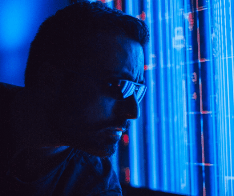 darkened image with blue tinge. White man looking at screen out of shot. In 40s wearing dark rimmed glasses blue lines behind to the side of him