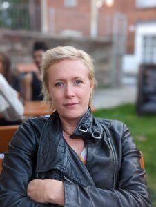 white woman very short blond hair black leather jacket looking at the camera blurred background 
