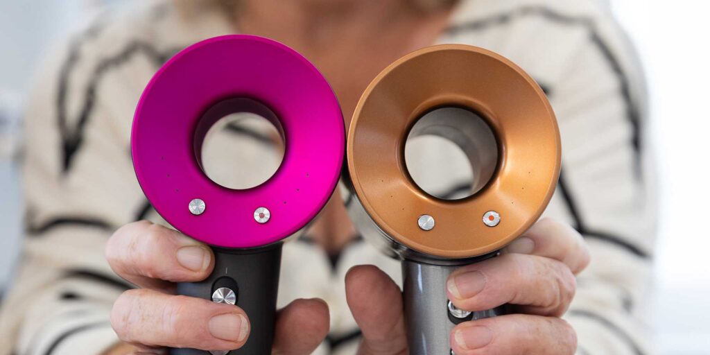 pair of hands holding out hair dryers one with bright pink edge one with gold