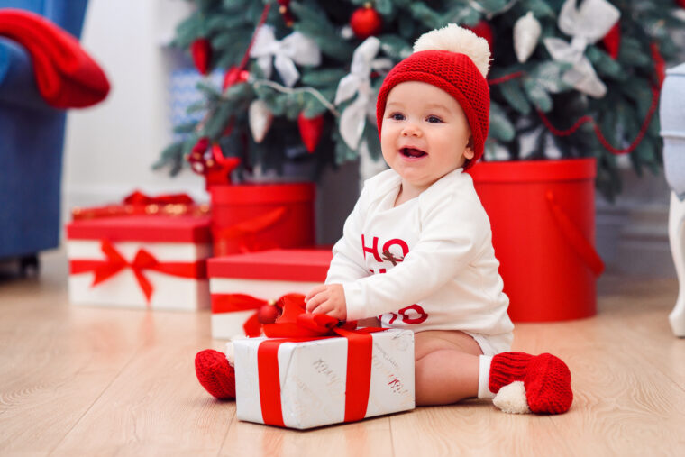 Toddler in white jumper Christmas red and white hat red booties in front of a Christmas tree with hands on box wrappen in white paper with red ribbon