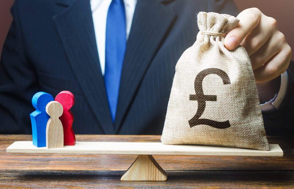 Man see just front of bule jacket of suit white shirt and blue tie puts pound sterling money bag on scales with a wooden piece family.