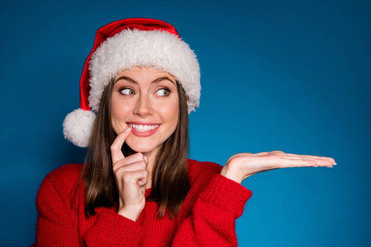 Photo positive girl in santa claus hat hold hand demonstrate x-mas newyear, advert promo touch finger teeth wear warm winter season clothes isolated blue gradient colour background