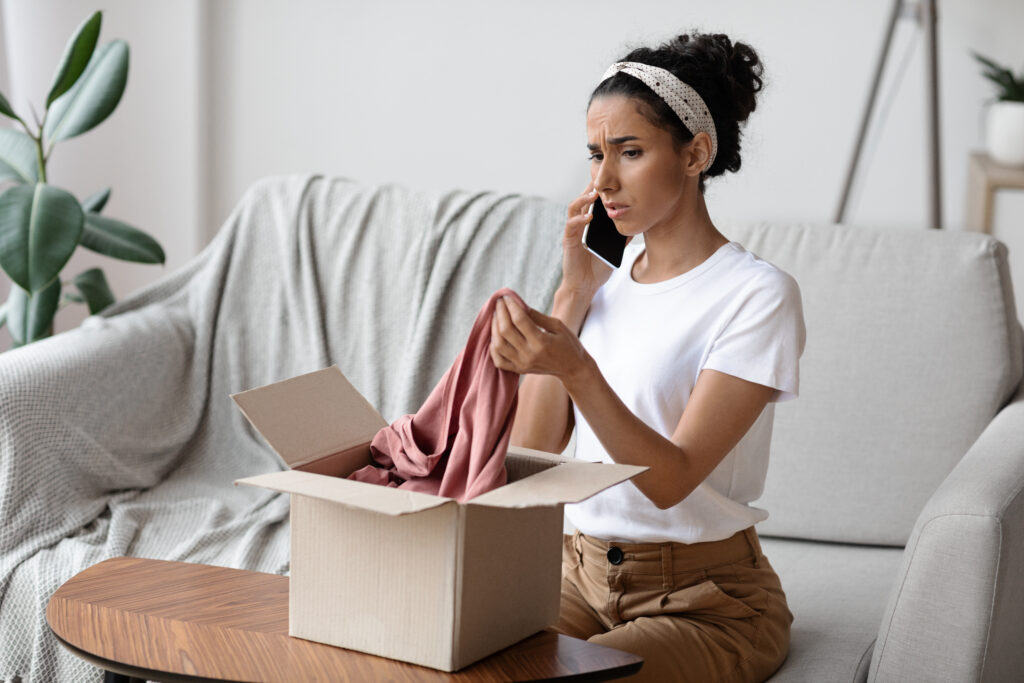 Disappointed young brunette woman sitting on sofa by wooden table and opening carton box, talking on mobile phone with seller after checking delivery, holding new clothes and complaining, empty space