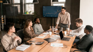Corporate Meeting Concept. Smiling Male Entrepreneur Giving Speech During Briefing With Coworkers Employees In Modern Office, Standing At Table In Board Room, Discussing Strategy And Management