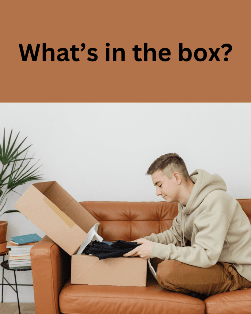 What's in the box title. White man in 20s sitting on brown sofa with grey hoodie on looking at open box holding a black jumper still in box