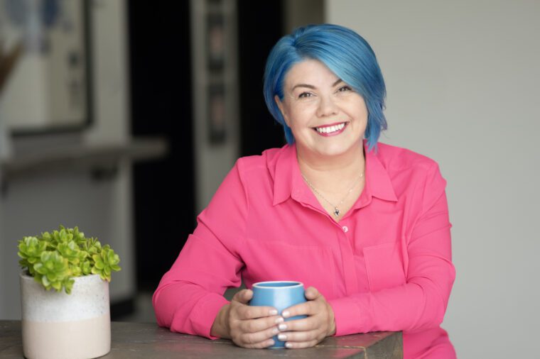Smiling white woman bright blue cropped hair, late thitries bright pink shirt hand round a mug on a table.