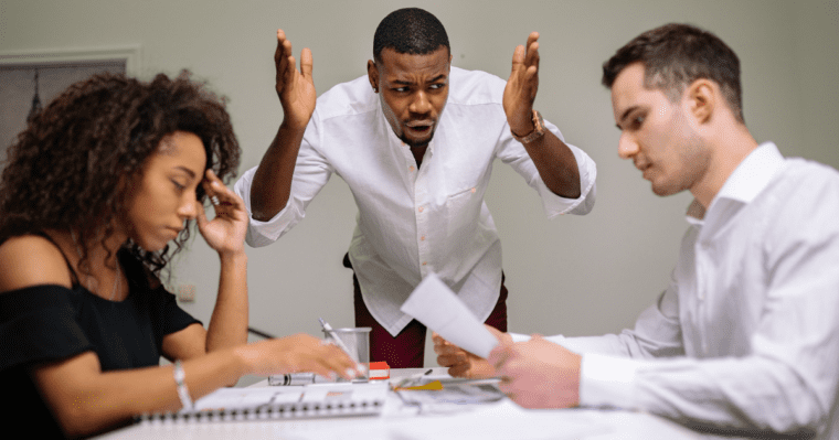 black woman sitting at a table looking down head in one hand at paper across from white man holding paper looking at it black man at end of table standing over with arms outstretched being ignored