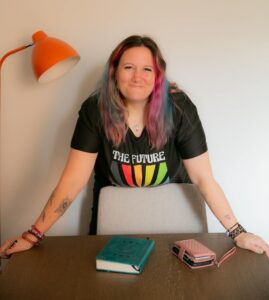 Maddy Alexander-Grout woman in 40s long rainbow coloured hair, smiling black t shirt arms outstretched on table with purse and wallet on the table. Orange angle poise light to the left