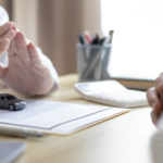 either side of table showing two arms and hand of people one over a document pot of pens to the side